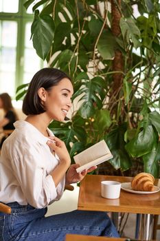 Vertical shot of asian brunette girl sitting elegant with book, reading in cafe and smiling, drinking coffee.