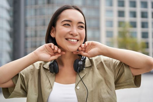 Portrait of smiling asian girl with headphones, posing in city centre, listening music.