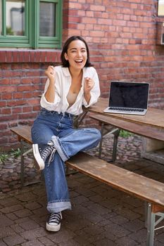 Vertical shot of enthusiastic asian girl, sits with laptop, screen is empty and blank, woman laughing and celebrating, triumphing.