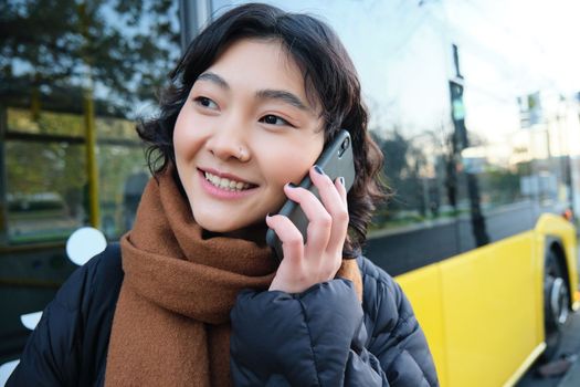 Cellular technology and people concept. Stylish asian girl talks on mobile phone, makes a telephone call, stands near bus stop and has conversation. Copy space