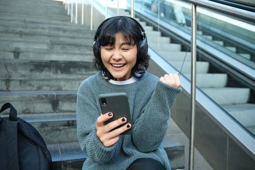 Enthusiastic korean girl in headphones, watches video and celebrates, looks at smartphone with triumphing, pleased face, say yes, sits on stairs in public place.