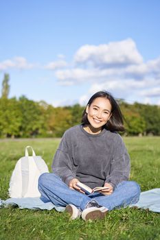 Vertical shot of cute teen girl sits in park on grass with backpack and her book, reading alone outdoors.