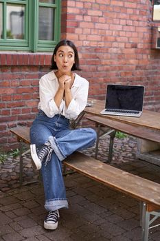 Full size portrait of asian girl sits with laptop, looks amazed and surprised and peeks at screen. Concept of people and urban lifestyle.