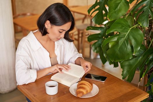 Beautiful young asian woman sitting in cafe with a book, eating croissant and reading, drinking cup of coffee. Wellbeing and self-care