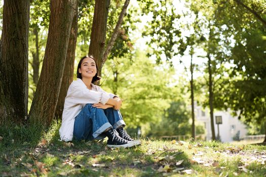 Portrait of beautiful asian woman resting near tree, relaxing in park, smiling and looking happy. Copy space