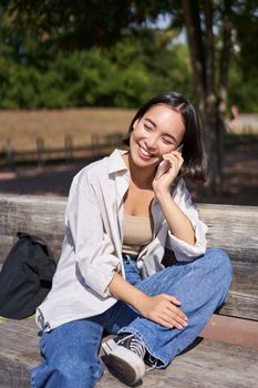 Smiling asian girl relaxing in park, talking on smartphone, having a mobile call while resting outdoors.