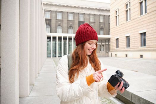 Smiling redhead girl photographer, taking pictures in city, makes photos outdoors on professional camera. Young talent and hobby concept