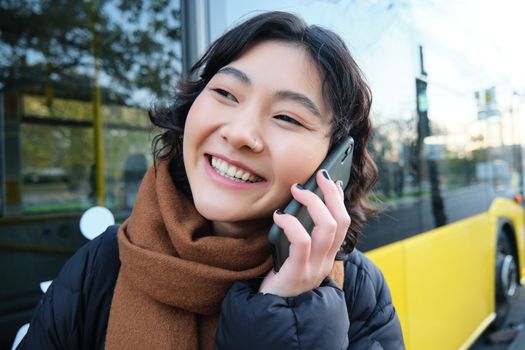 Close up portrait of happy asian girl talks on mobile phone, smiles while speaks to someone on smartphone, stands near bus on stop.