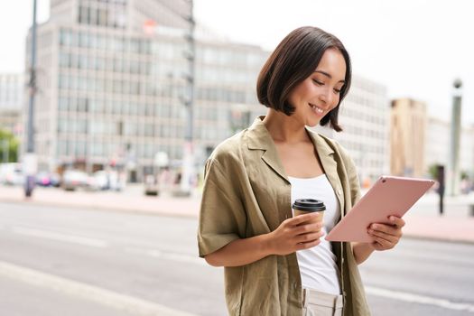 Happy city girl walking on streets, using tablet and drinking coffee on her way to work.