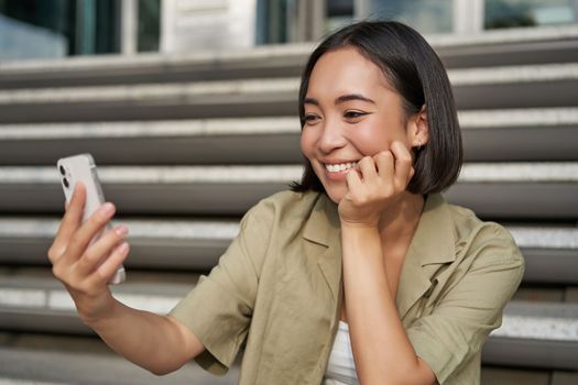 Portrait of happy smiling asian girl takes selfie on mobile phone. Young korean woman video chat on smartphone while sitting on stairs near building.