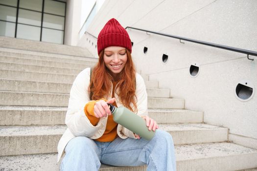 Happy redhead woman, tourist with thermos, drinks her hot tea, coffee from travel flask, restests during her travelling in city and sightseeing.