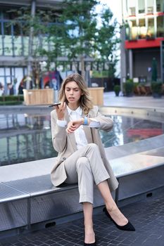 Vertical shot of smiling woman records voice message and looks at digital watch, reads message or checks time, sits in city centre.