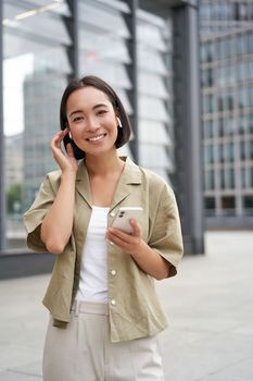 Young smiling asian woman in wireless earphones, walks on street with smartphone and headphones.