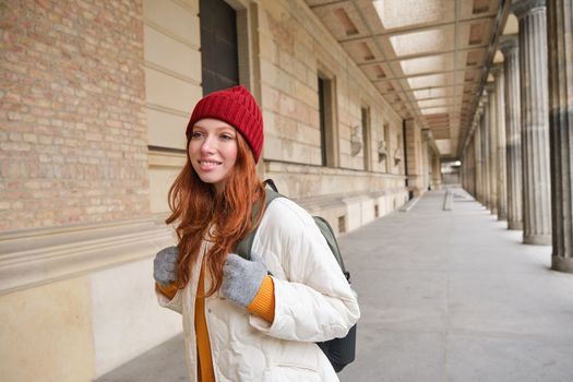 Smiling redhead girl with backpack, walks in city and does sightseeing, explore popular landmarks on her tourist journey around Europe.