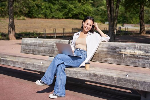 Portrait of beautiful young woman sitting on bench in park, using laptop and drinking coffee, relaxing outdoors on summer day.