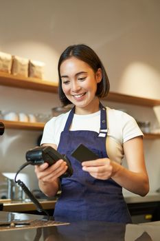 Portrait of smiling asian barista, coffee shop employee using POS terminal and credit card, helps client pay contactless in cafe.