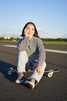 Vertical shot of asian girl skater, sits on her skateboard and smiles, enjoys sunny day, cruising on longboard on empty road outdoors.