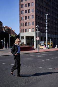 Vertical portrait of confident and stylish businesswoman in suit, walking, cross the street in city center, wears sunglasses.