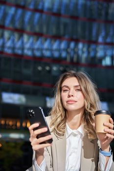 Stylish young corporate woman standing in street, drinks coffee and holds mobile phone.