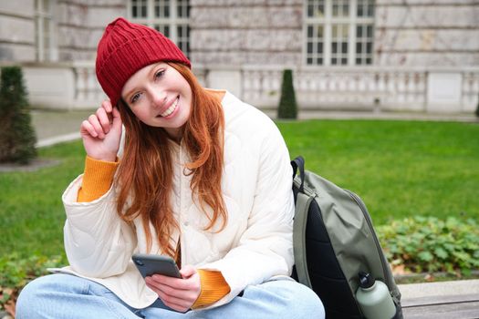 Young smiling redhead girl sits in park with smartphone, texting message on mobile phone, using telephone, connecting to public wifi and surfing internet.