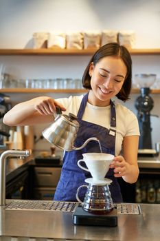 Vertical shot of smiling asian bartender, barista in blue apron, pouring water with small kettle, brewing coffee behind counter in her cafe.