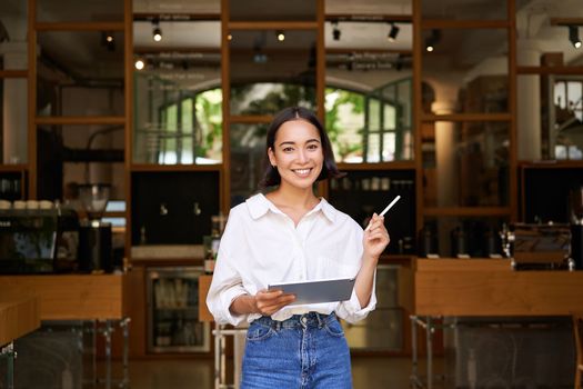 Portrait of asian woman, manager standing with tablet in front of cafe entrance, welcomes guests. Business and entrepreneurs concept