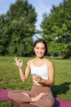 Vertical shot of asian woman shows okay sign, recommending yoga training online, meditation app, doing exercises on fresh air in park.