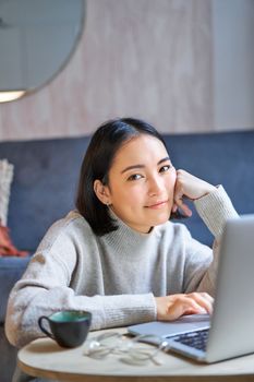 Vertical shot of young asian woman looking tired, smiling with exhausted expression, working from home on her laptop.