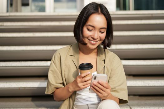 Happy urban girl drinks her takeaway coffee and scrolls feed on smartphone. Asian woman sits on stairs with tea and holding mobile phone.