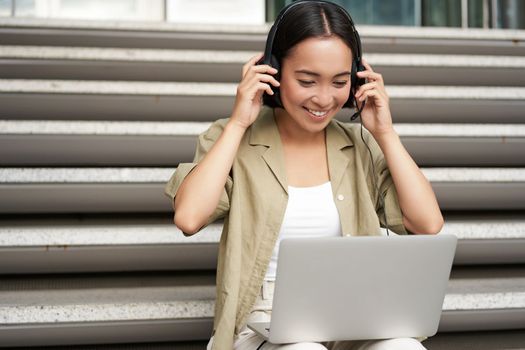 Portrait of asian woman, digital nomad girl using laptop and listens to music outdoors. Young student works on computer and smiles.