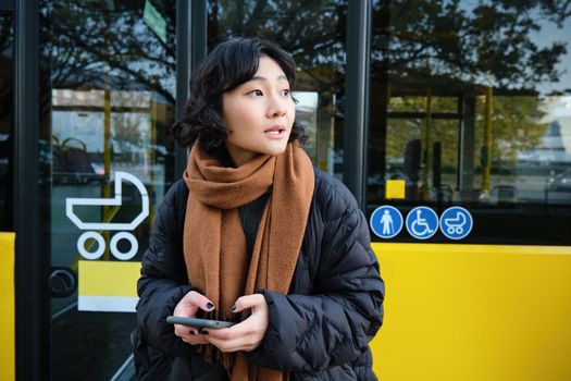 Portrait of korean girl looking for her bus on a stop, holding mobile phone, checking schedule, time table on smartphone app.