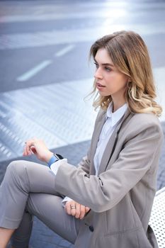 Vertical shot of successful corporate woman in suit, checking time on watch, looking away, waiting for someone in city, sitting outside.