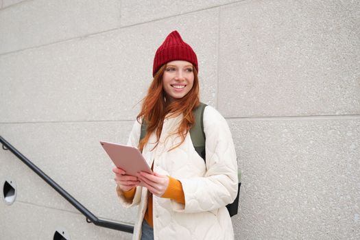 Young redhead girl, walks with digital tablet around city, stands on street with backpack and gadget, looks at online map, searches for a place in internet.