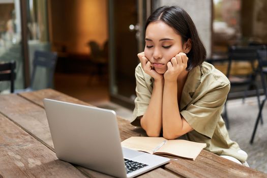 Bored girl looks at her laptop, sits outdoors in cafe, listening boring online meeting, working and feeling sad.