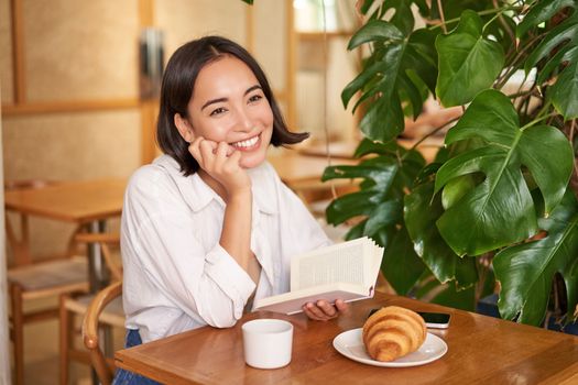 Romantic asian woman sitting with book in cafe, eating croissant and drinking coffee, reading and smiling, enjoying alone time.