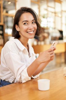 Vertical portrait of stylish asian woman sitting in cafe, drinking coffee and using smartphone. Lifestyle and people concept