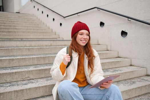 Happy redhead woman in red hat, sits on stairs outdoors, uses tablet, connects to wifi near public building, reads digital book while waits on street.
