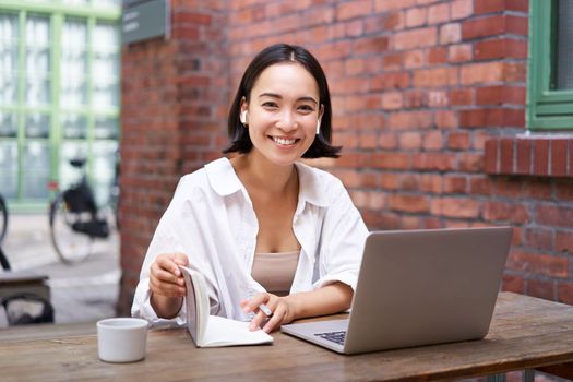 Stylish asian girl works in coworking with laptop, makes notes, uses wireless earphones and smiles, sits near brick wall in office.