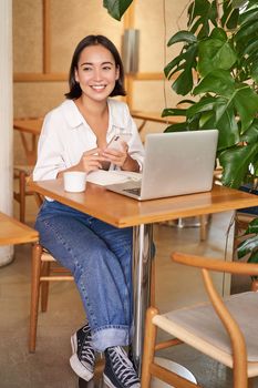 Vertical shot of young asian girl sits in cafe with laptop and smartphone, relaxing and surfing the net, working remotely.