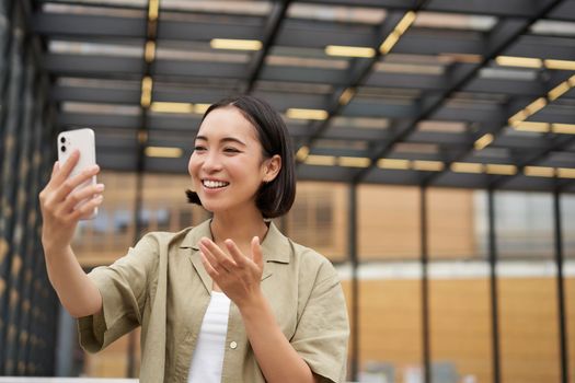 Happy asian girl shows something behind her during video call, demonstrating smth, standing on street.