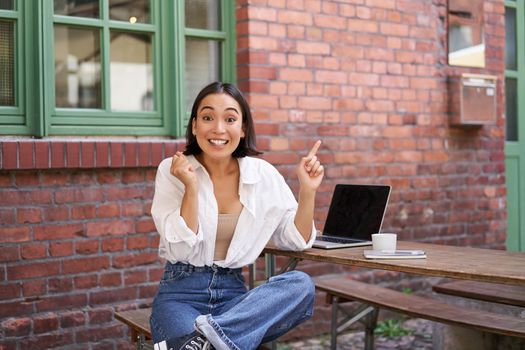 Stylish urban girl, asian woman with laptop sits in cafe, points at upper right corner banner, shows copy space with surprised face expression.