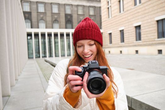 Smiling redhead girl photographer, taking pictures in city, makes photos outdoors on professional camera. Young talent and hobby concept