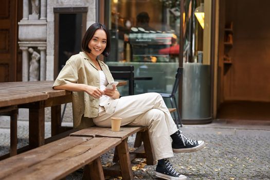 Portrait of stylish young korean woman sits in cafe, holds smartphone, smiles, enjoys coffee outdoors.