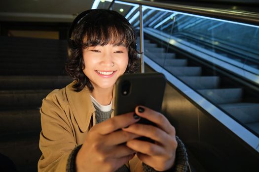 Portrait of happy asian girl in headphones, smiling and looking at smartphone, watching video or listening music on mobile phone, sitting on staircase.