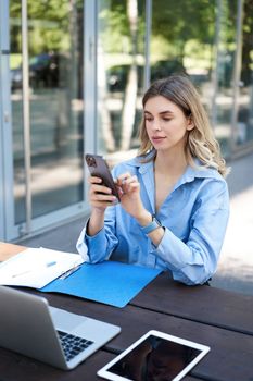 Portrait of businesswoman working outside in park, sitting with laptop, work documents and using mobile phone, text message, reading on smartphone.