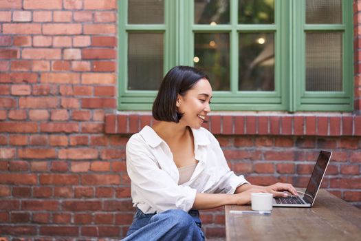 Portrait of young stylish woman, influencer sitting in cafe with cup of coffee and laptop, smiling and looking confident.