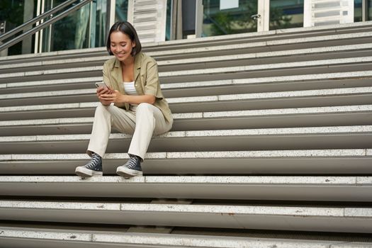 Portrait of smiling asian girl sits on stairs outdoors, sending message, using smartphone app, looking at mobile phone screen.