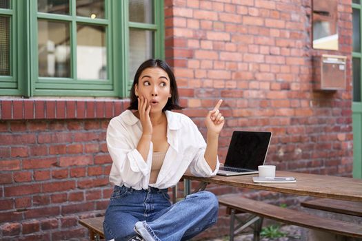 Stylish urban girl, asian woman with laptop sits in cafe, points at upper right corner banner, shows copy space with surprised face expression.