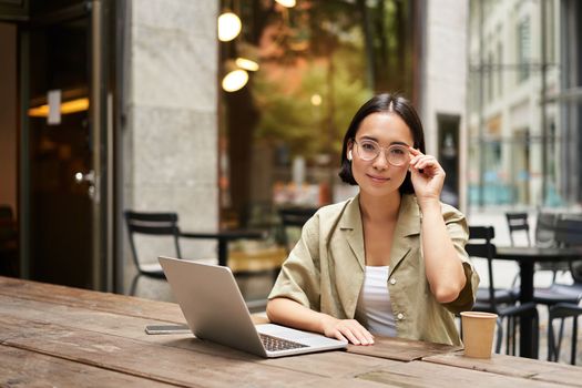 Young woman, student working from cafe, sitting with laptop and cup of coffee outdoors, looking at camera.