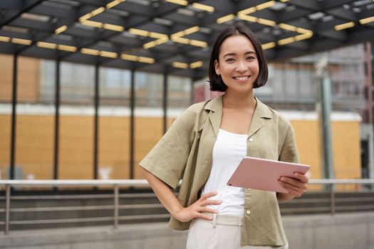 City girl standing with tablet on street, using application, smiling at camera.